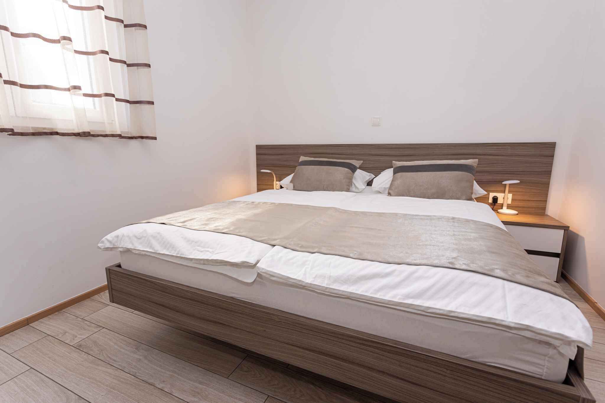 Una cama bien hecha con un cabecero de madera, dos mesitas de noche con lámparas y una ventana con cortinas translúcidas.
