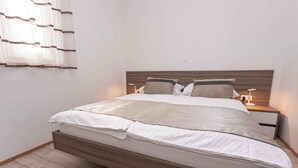 Una cama bien hecha con un cabecero de madera, dos mesitas de noche con lámparas y una ventana con cortinas translúcidas.