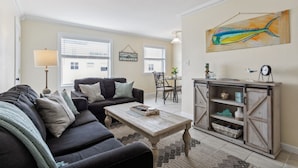 A living room with a dark sofa, a wooden coffee table, a sideboard with open shelves, and a painting of a fish on the wall. - 1 block to beach & boardwalk | Central Downtown (Virginia Beach)