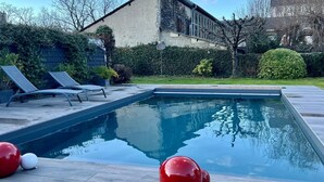 Een rechthoekig zwembad met een helder blauw wateroppervlak, omringd door een houten terras. Er staan twee ligbedden en drie rode, bolvormige objecten op het terras.