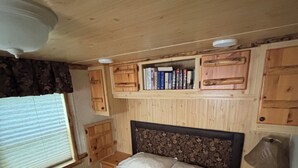 Un dormitorio con cama, cabecero de madera, estanterías empotradas con libros, mesita de noche con lámpara y ventana con persianas.