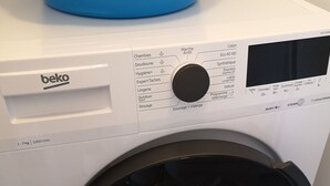 Voorweergave van een witte beko wasmachine met een zwarte deur, een bedieningspaneel met diverse instellingen, en een blauwe bak bovenop.