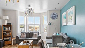 A modern living room with a sofa, a coffee table, a bookshelf, a dining area with a glass table, and a view of the sea through large windows. - The Penthouse, 16 At The Beach, Wi-Fi, Sea View (Kingsbridge)