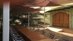 An outdoor dining area with a long wooden table and metal chairs, shaded by a large umbrella, set against a backdrop of greenery and a stone wall with a wooden door. - Still available from August 17 to August 31! La Xaconera d' Ardenya (Ardenya, Tarragona)