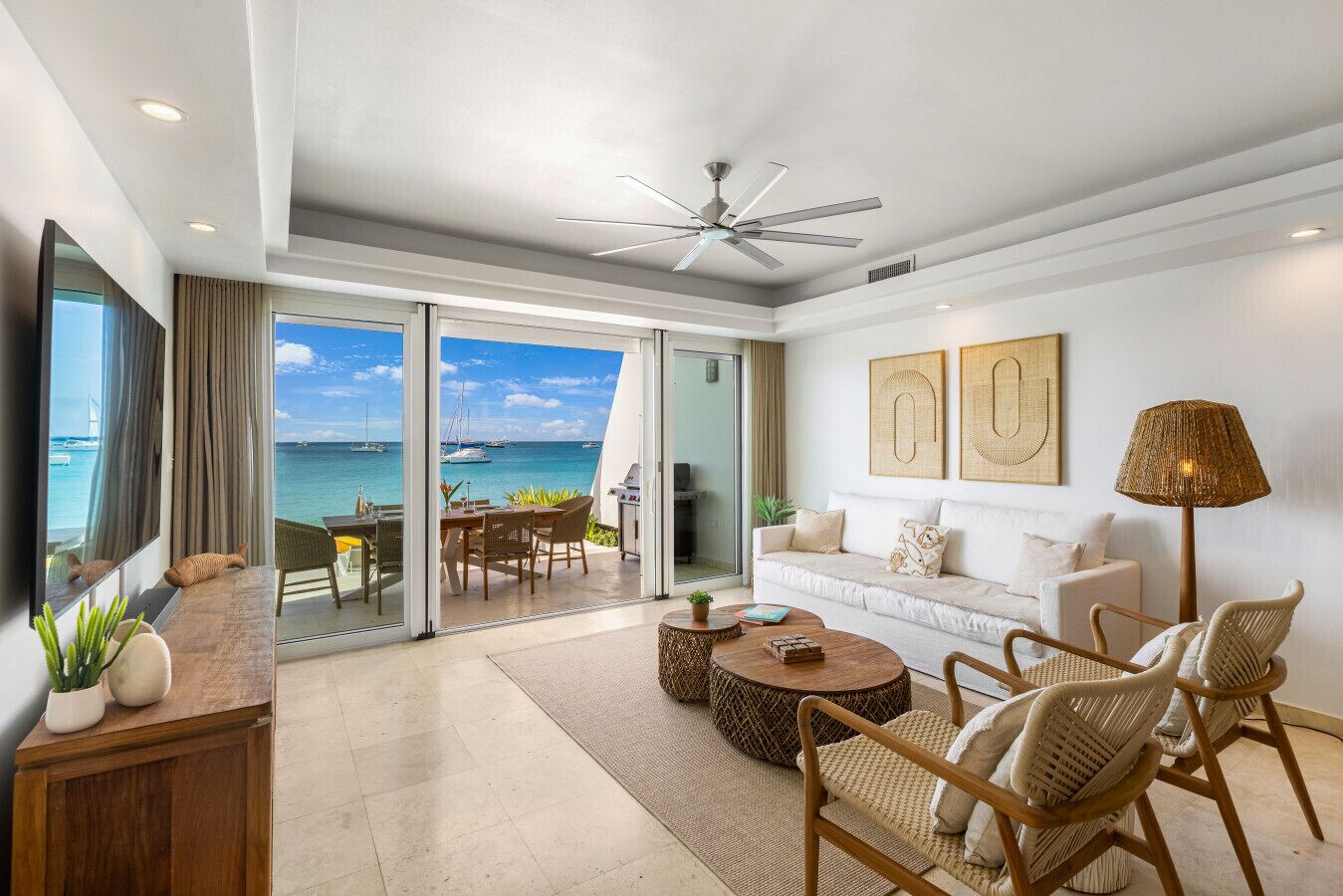 A spacious living room with a view of the ocean, a ceiling fan, and a large flat-screen TV.