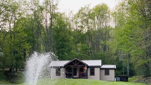 A lakeside view with a fountain, a small building with a porch, and a grassy area. - Charming cottage with pond in Heart of New River Gorge (Victor)