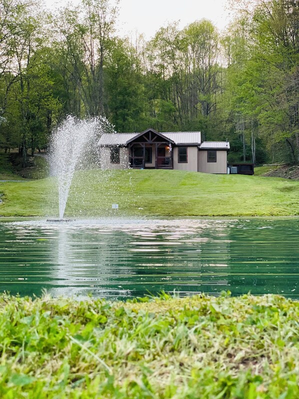 A lakeside view with a fountain, a small building with a porch, and a grassy area. - Charming cottage with pond in Heart of New River Gorge (Victor)