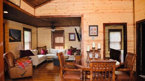 A cozy log cabin interior with a dining area, a sofa, and a fireplace. - Charming cottage with pond in Heart of New River Gorge (Victor)