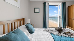 A bedroom with a bed, a bedside table, a lamp, a framed picture, a window with curtains, and a view of the beach. - 11 At The Beach, Torcross - an apartment that sleeps 4 guests in 2 bedrooms (Kingsbridge)