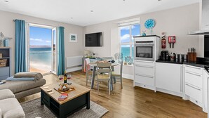 A modern living room with a sofa, a coffee table, and a dining area with chairs. A kitchen with a stove and cabinets is visible. - 11 At The Beach, Torcross - an apartment that sleeps 4 guests in 2 bedrooms (Kingsbridge)
