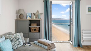 A room with a grey cabinet, a sofa with patterned cushions, a window with blue curtains, and a view of a beach. - 11 At The Beach, Torcross - an apartment that sleeps 4 guests in 2 bedrooms (Kingsbridge)