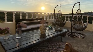 Een houten tafel met een vaas, een fles en een kussen op een balkon met een witte balustrade.