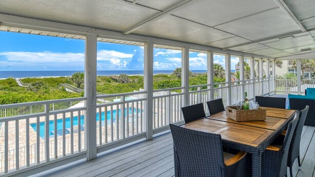Ein überdachter Außenbereich mit Holztisch und -stühlen, einem Pool und Blick auf Meer und Grünflächen.