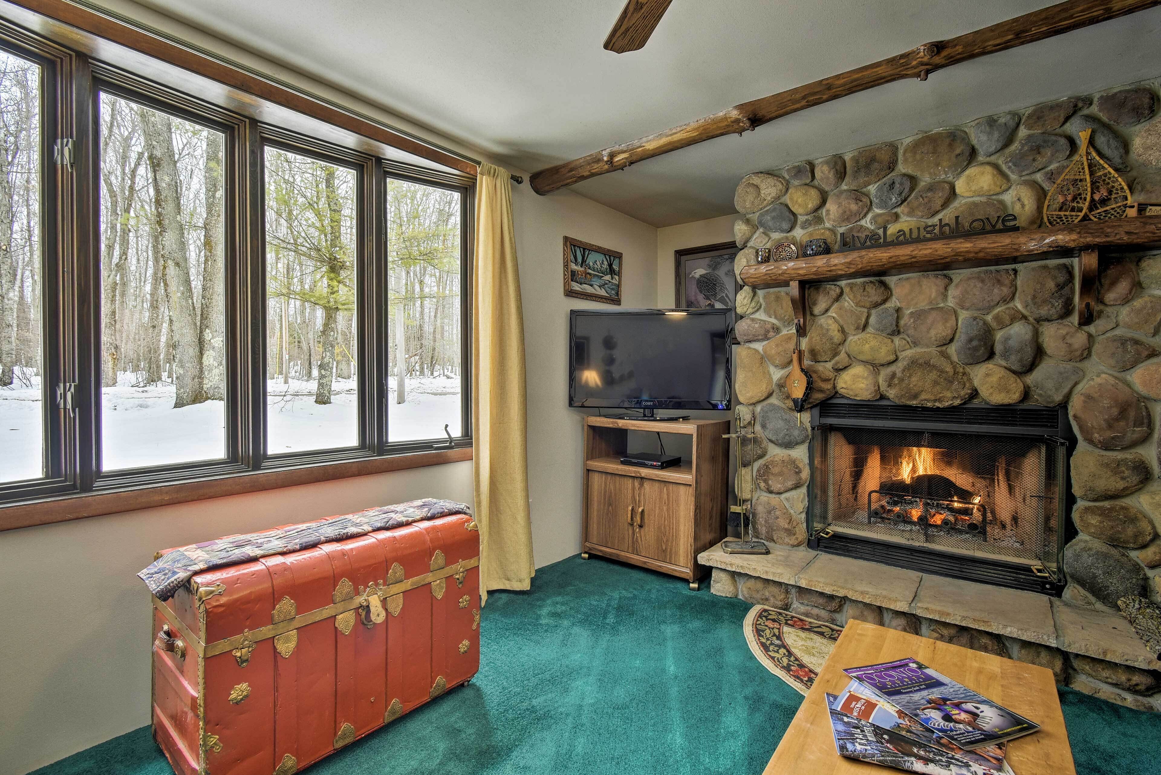 Una sala de estar acogedora con chimenea de piedra, un baúl rojo, televisión y vistas a un bosque nevado a través de amplios ventanales.