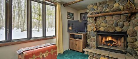 Una sala de estar acogedora con chimenea de piedra, un baúl rojo, televisión y vistas a un bosque nevado a través de amplios ventanales.