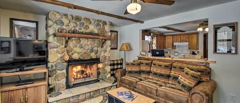 A cozy living room with a stone fireplace, a ceiling fan, a large sectional sofa, and a television.