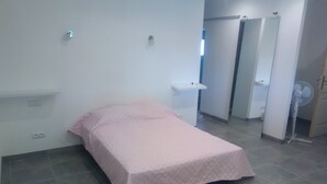 A small, neatly arranged bedroom with a single bed, a bedside table, a small white shelf, and a wall-mounted fan.