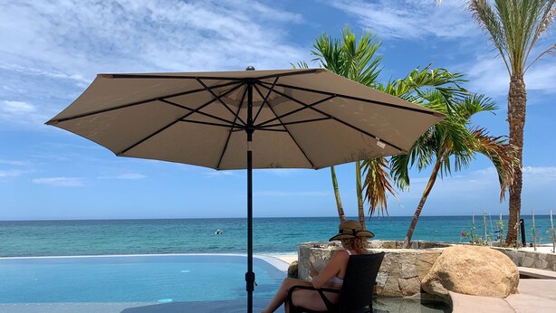 En person sidder under en strandparasol ved en pool med udsigt til havet.