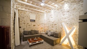 A cozy living room with a stone wall, a wooden ceiling, a sectional sofa, a coffee table, and a large 'X' light fixture. - Alice Residence - Two Storey Luxury Residence in the Heart of Rethymno Old Town (Rethymno)