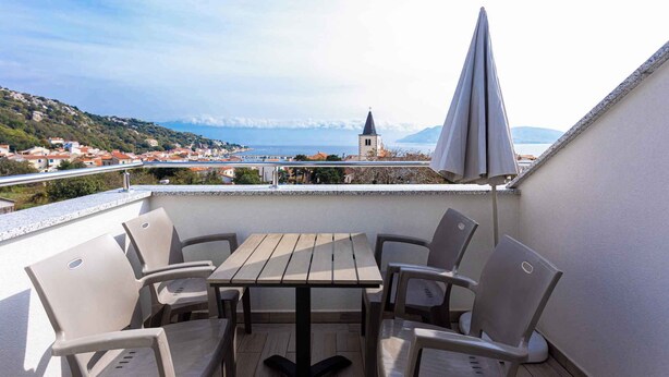 A balcony with a table and chairs, overlooking a coastal town with a church and mountains in the distance. - Holiday apartment mit Klimaanlage und Meerblick (Baška)