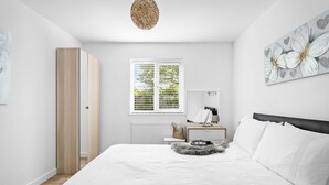 Een slaapkamer met een bed, nachtkastje, lamp, raam met zonwering en een bloemendecoratie op de muur.