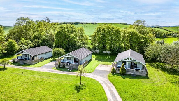 Een klein, tweegelegen huis met terras en hottub, omringd door weelderige groene velden en bomen.