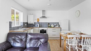 Een compacte keuken met moderne apparaten, een eethoek met een houten tafel en witte stoelen, en een leren bank in de woonkamer.