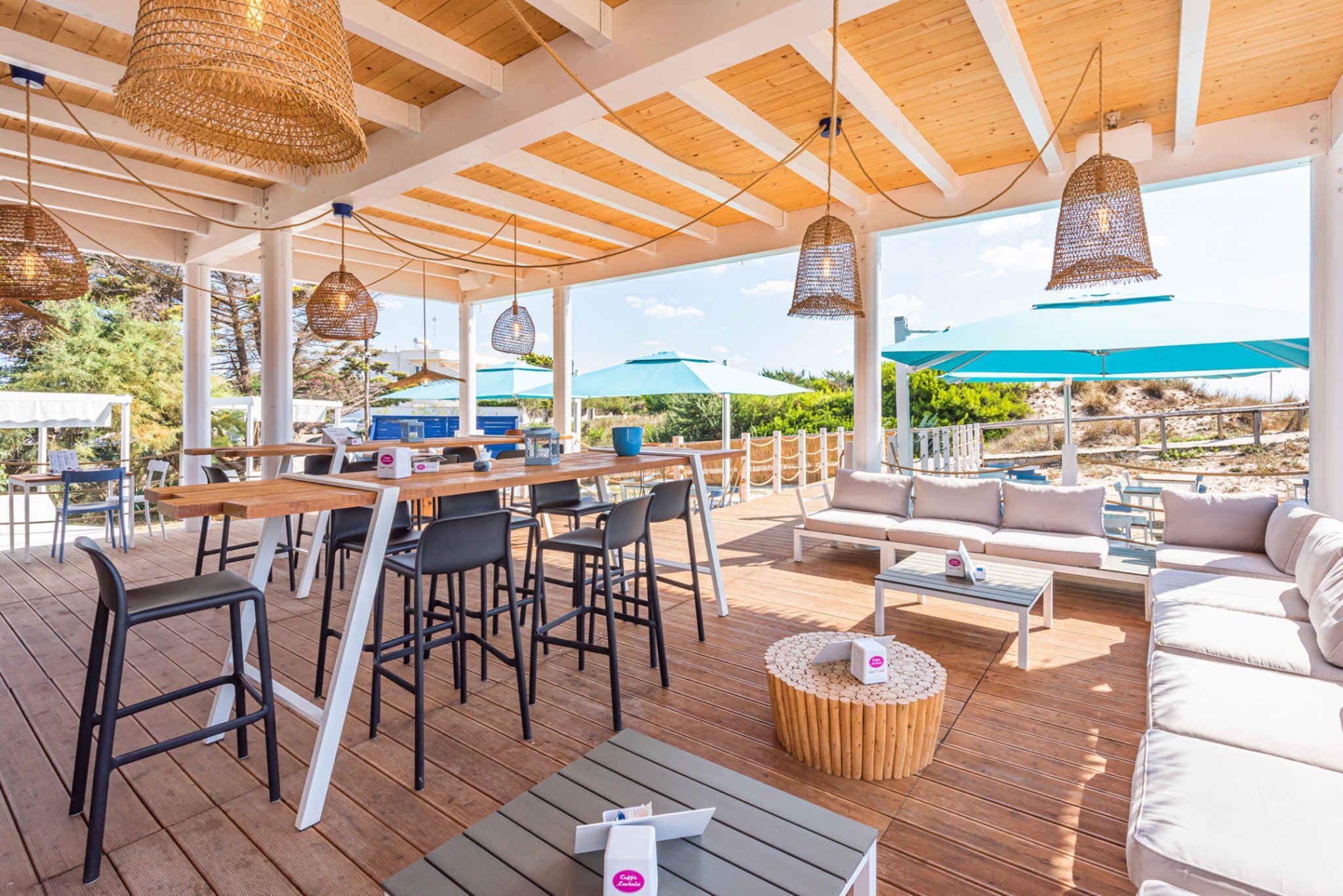 Un patio al aire libre con mesas, sillas y un sofá de madera, con lámparas colgantes de tejido y vistas al paisaje.