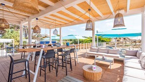 Une terrasse extérieure avec des tables, des chaises et un canapé en bois, agrémentée de luminaires suspendus en fibres tressées et offrant une vue sur le paysage.