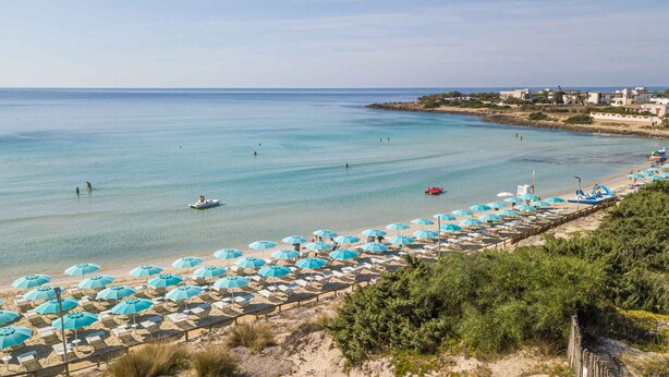 Une plage bordée de rangées de parasols et de chaises longues, une mer d’un bleu limpide et des bâtiments en arrière-plan.