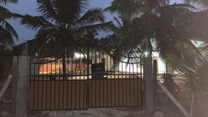 A gated compound with a metal gate, palm trees, and a building with a red roof in the background.