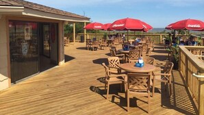 Een terras buiten met houten tafels en stoelen, rode parasols en uitzicht op de oceaan.
