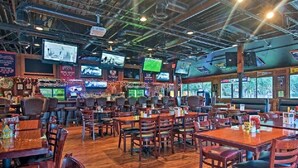 Een ruim restaurant met meerdere tafels en stoelen, een bar en verschillende televisies aan de muur.