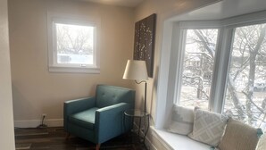 A room with a blue armchair, a lamp, a window seat with cushions, and a view of trees. - Cozy 1-bedroom condo in downtown Saugatuck, perfect for a romantic getaway. (Saugatuck)
