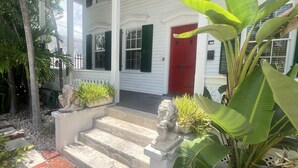 Un edificio blanco de dos pisos con contraventanas verdes, una puerta roja y un porche con escalones.