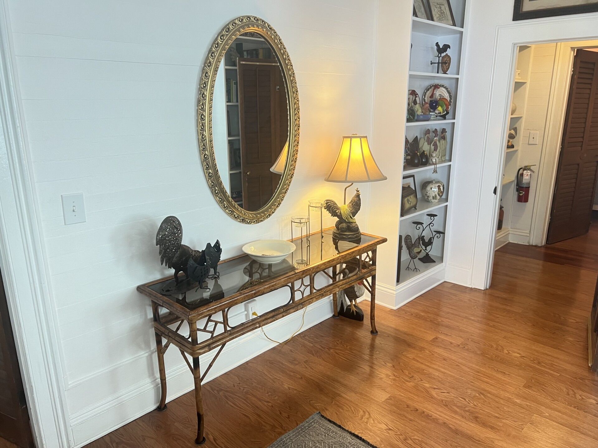Una consola de madera con tapa de cristal, un espejo con marco dorado, una lámpara y objetos decorativos, incluyendo una estatua de gallo.