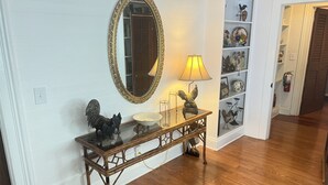 Una consola de madera con tapa de cristal, un espejo con marco dorado, una lámpara y objetos decorativos, incluyendo una estatua de gallo.
