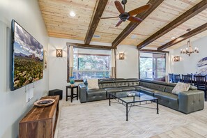 Un salón espacioso con un sofá modular grande, una mesa de centro de vidrio y un ventilador de techo. Hay dos ventanas que ofrecen luz natural y vistas al exterior.