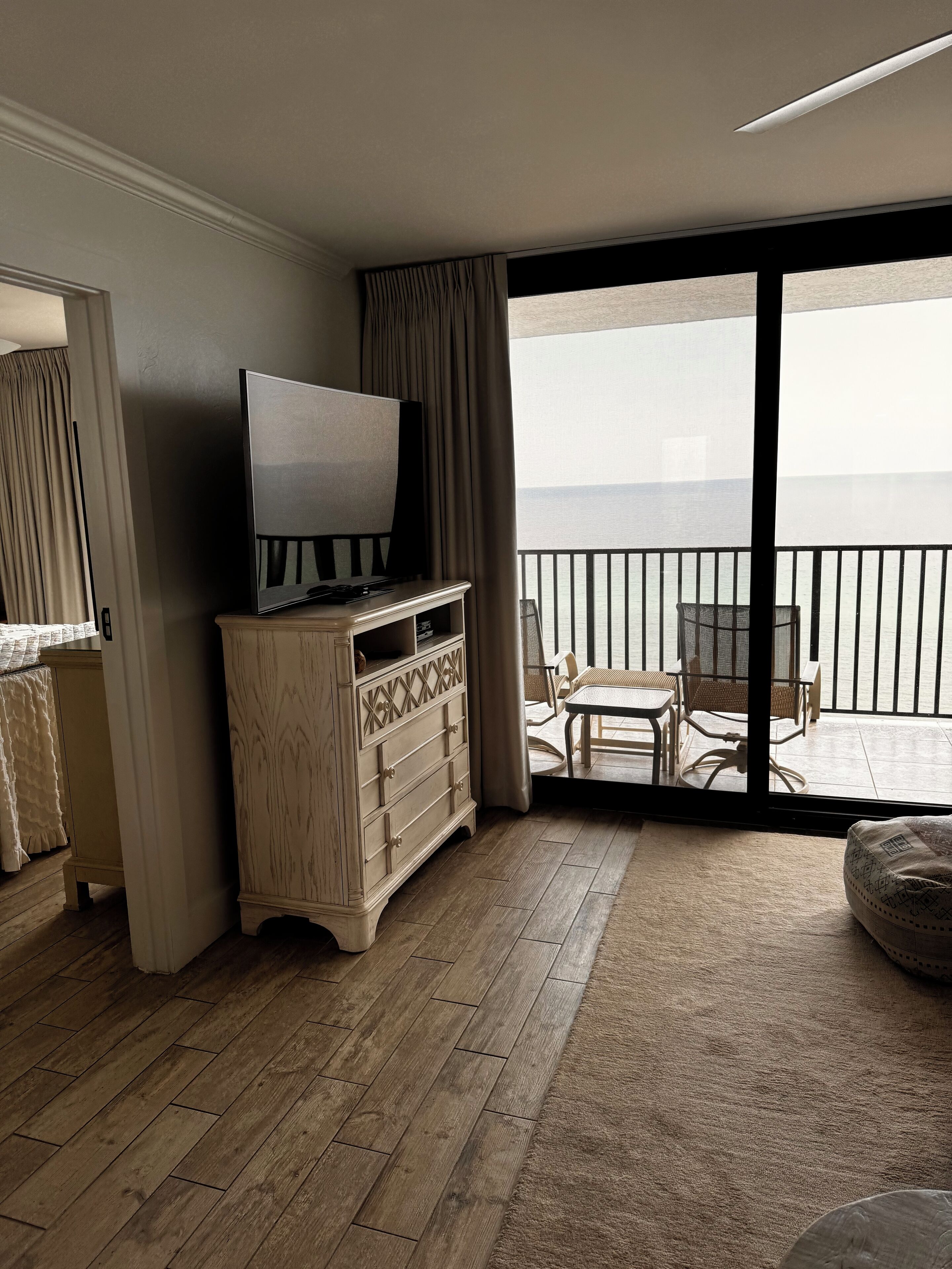 Een hotelkamer met een houten dressoir, een televisie, een balkon met uitzicht op zee en een zithoek met stoelen en een tafel.