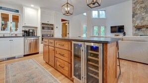 A modern kitchen with white cabinets, stainless steel appliances, and a central island. There are two pendant lights, a stone accent wall, and a flat-screen TV mounted above the island.
