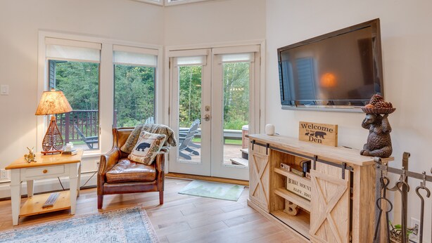 A cozy living room with a leather sofa, a wooden coffee table, and a TV mounted on the wall. There are large windows providing a view of the outdoors.