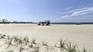 Una playa con una sombrilla azul y una silla, una zona arenosa con césped, y un cielo despejado.