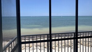 Blick von einem Balkon mit Metallgeländer auf einen Strand mit klarem Wasser und weiter entfernte Boote.