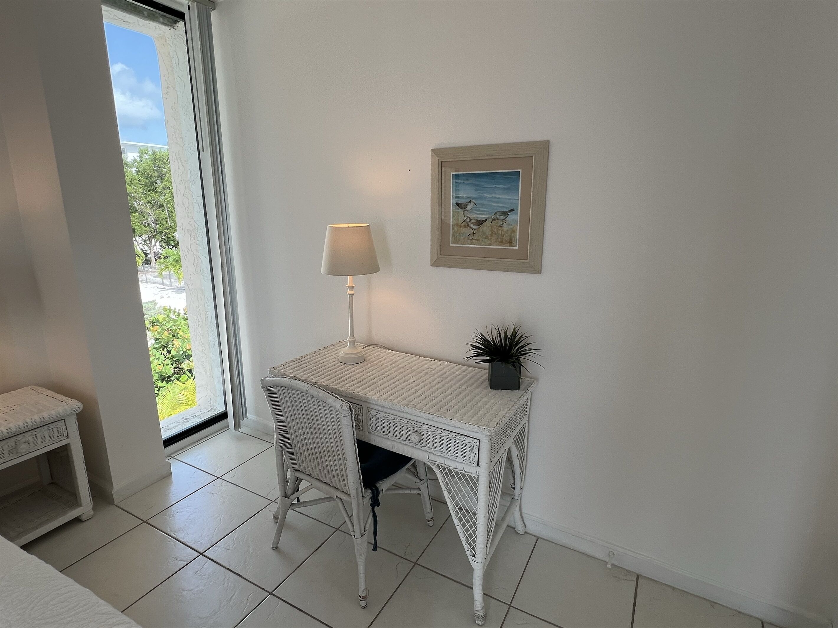 Un bureau en osier blanc, avec une lampe et un tableau encadré accroché au mur.