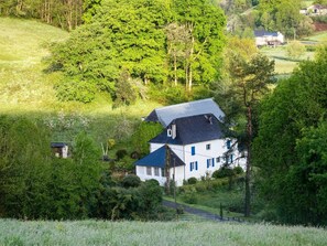 A white house with a dark roof, surrounded by lush greenery and trees.