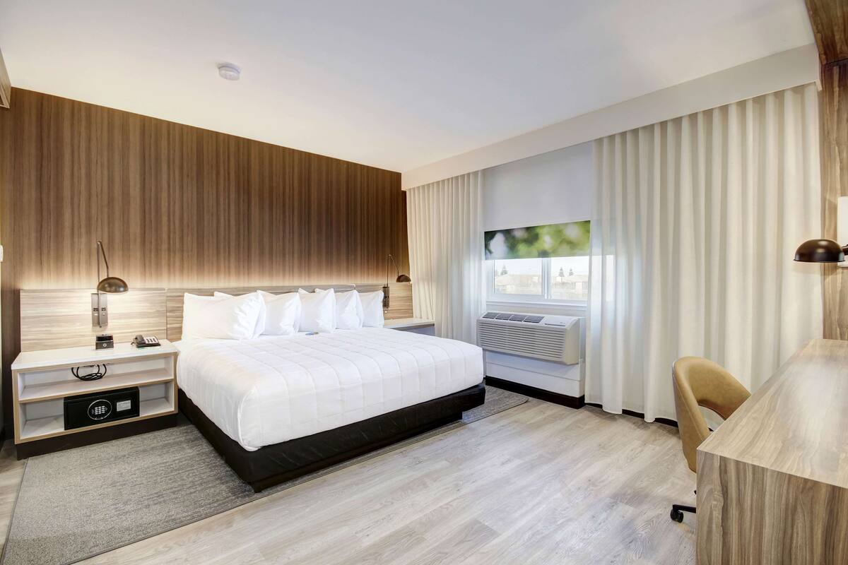 A modern hotel room with a large bed, bedside tables, a desk, and a view of the outdoors through the window.