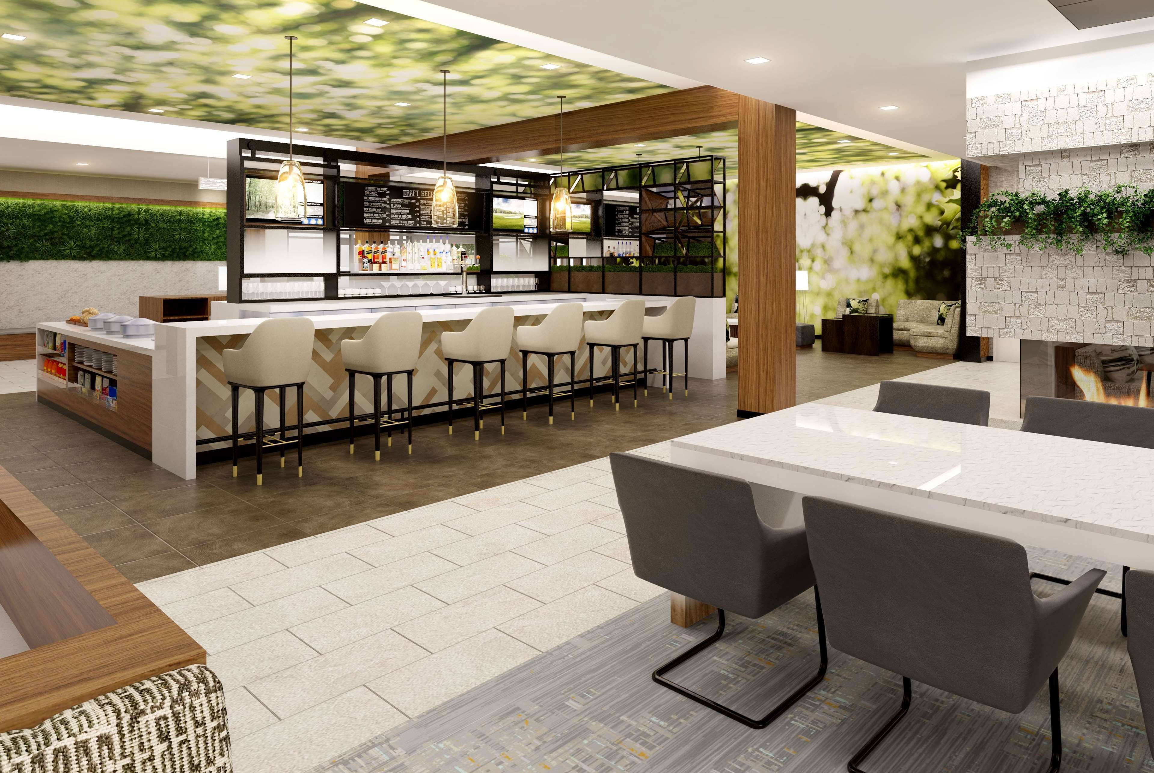 Un área de bar moderna con una barra alargada y varios taburetes, una zona de estar con una mesa y sillas, y una pared decorativa con follaje.