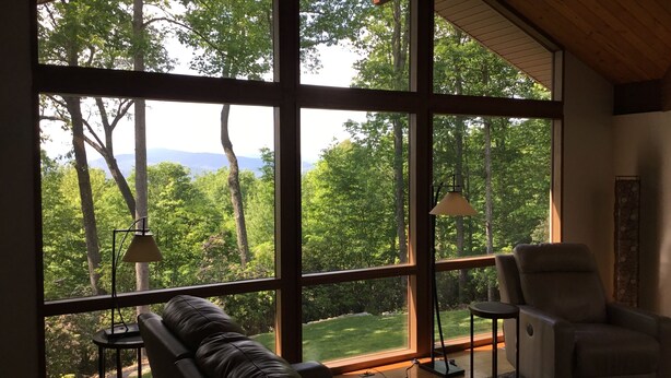 A room with large windows offering a view of a forest and mountains. There is a leather sofa and a chair. - 2.8 Acres of Mountain Magic.  Close to Everything.  (Pisgah Forest)