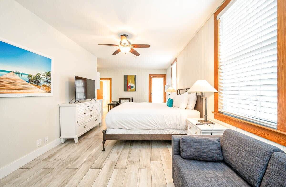 A bedroom with a bed, a sofa, a TV, a ceiling fan, and a window with blinds.