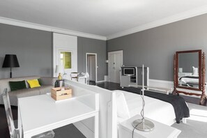 Una habitación de hotel moderna con un escritorio blanco, un sofá con cojines verdes, una mesita de noche con una lámpara, un televisor y un espejo grande.